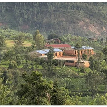 Load image into Gallery viewer, Rural landscape with brick buildings and lush green vegetation in a hilly region.
